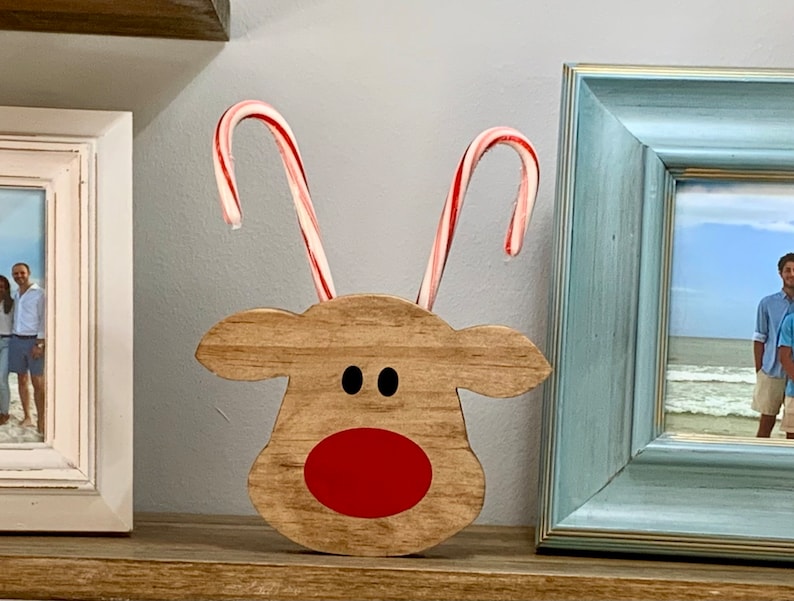 May include: A wooden reindeer figurine with a red nose and black eyes. The reindeer has two candy cane antlers.