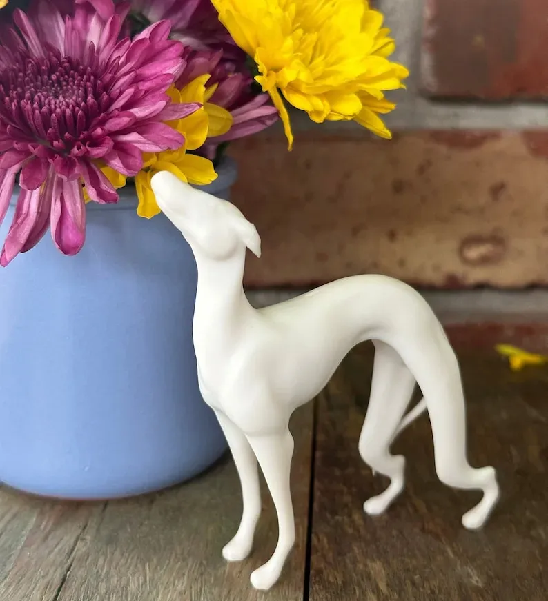 May include: A white ceramic figurine of a dog standing on a wooden surface. The dog is facing the left side of the image and has its head tilted up. The dog is standing in front of a blue vase with pink and yellow flowers.