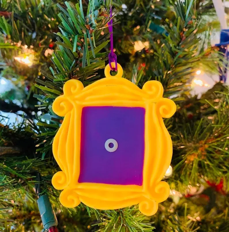 May include: A yellow plastic frame ornament with a purple center. The ornament is shaped like the iconic frame from the TV show Friends. The ornament has a string loop for hanging on a Christmas tree.