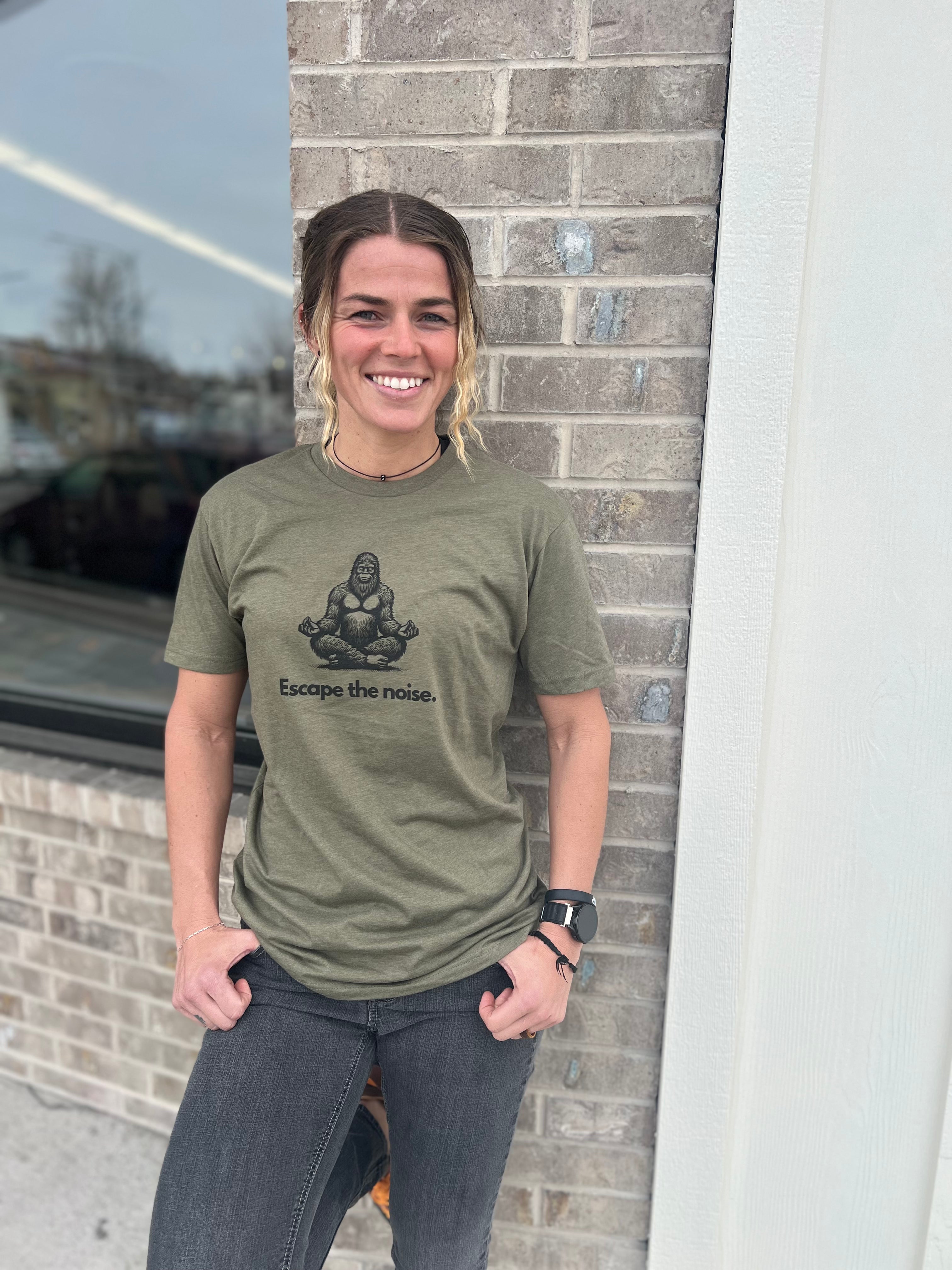 Woman wearing a Heather Military Green T-shirt with a Bigfoot graphic in a meditative pose and the text "Escape the noise," standing outside against a brick wall, showcasing Timpanogos Hiking Co.'s outdoor apparel.