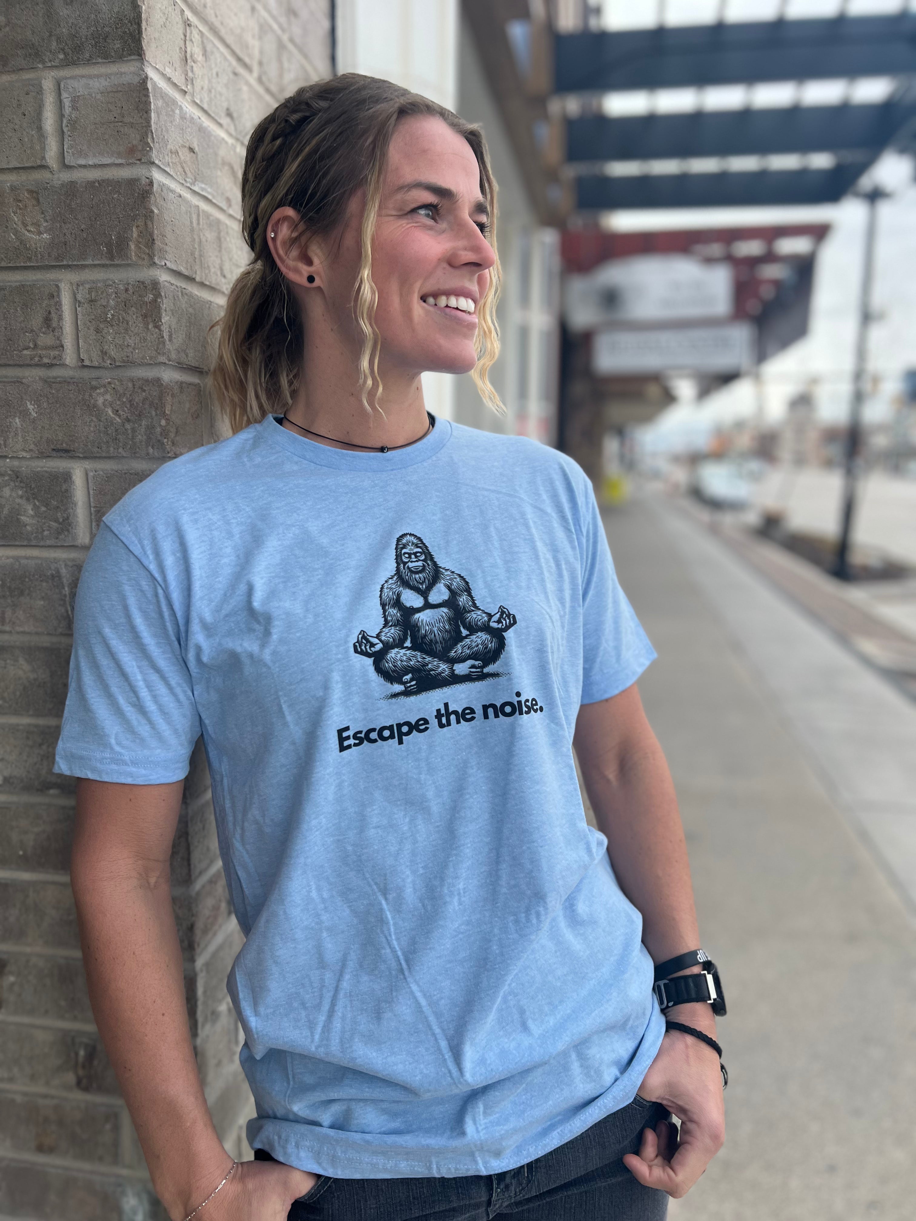 Woman wearing a Heather Blue T-shirt with a Bigfoot graphic in a meditative pose and the text "Escape the noise," standing outside against a brick wall with a city street backdrop, representing Timpanogos Hiking Co.