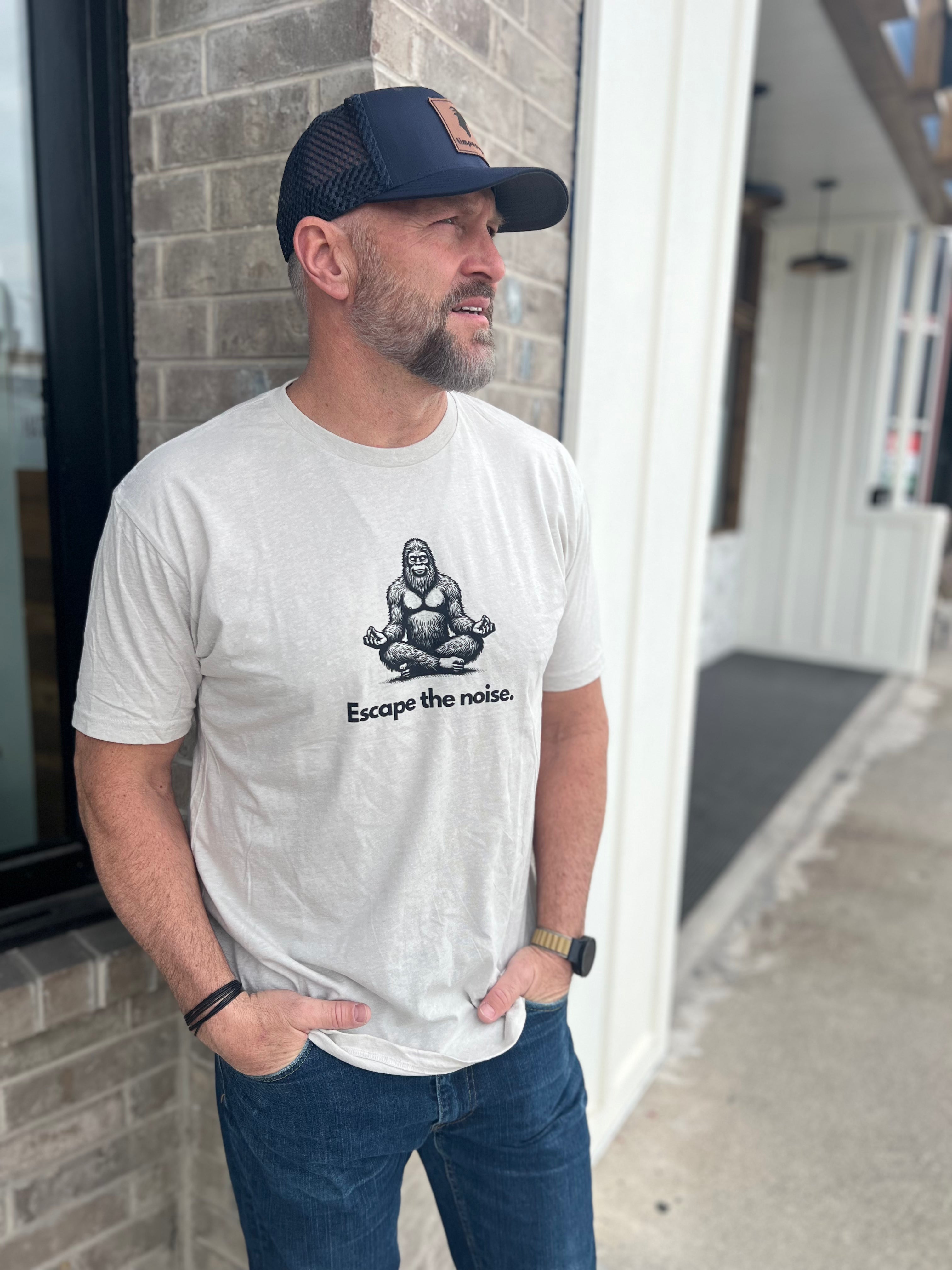 Man wearing a Heather Cream T-shirt with a Bigfoot graphic in a meditative pose and the text "Escape the noise," paired with a navy and black trucker hat, standing outside against a brick wall, showcasing Timpanogos Hiking Co. style.
