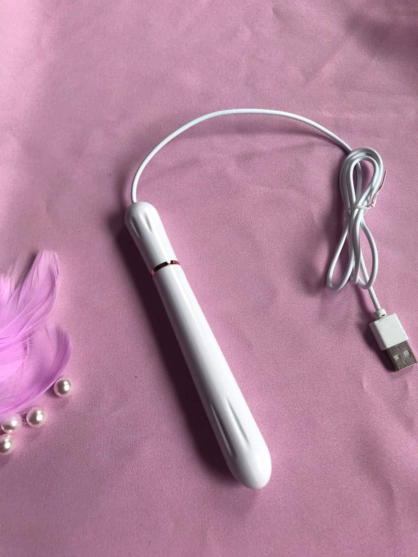 Lilysuck USB Heating Rod with white elongated body, gold-colored band, connected USB cable on soft pink fabric with white pearls and pink feathers