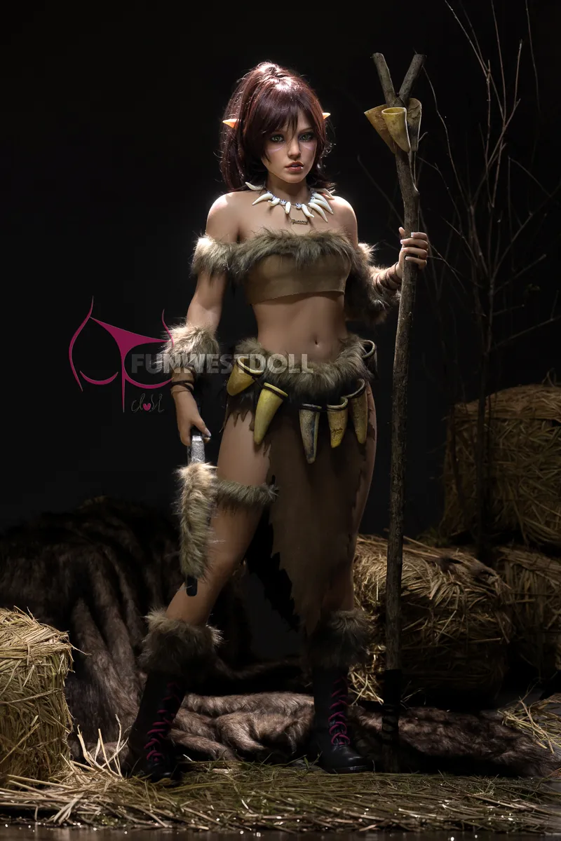 Realistic tribal warrior doll in fur outfit, holding a spear, standing on hay with dark background.