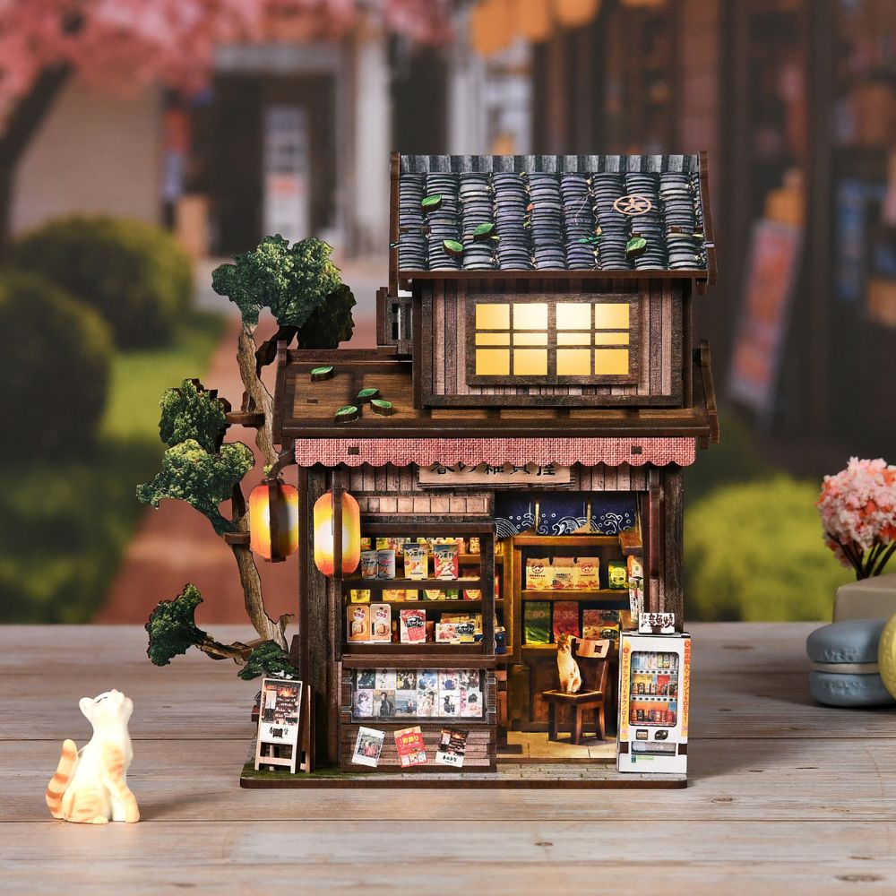 It is a detailed scale model of a traditional Japanese street scene, possibly a sweet shop or general store. Key features include a tiled roof, paper lanterns, a cat figure, and a miniature vending machine. 
