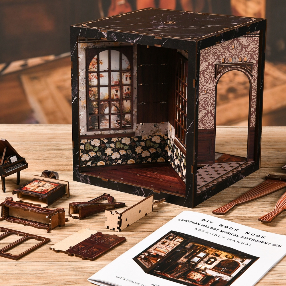 The miniature DIY book nook kit in progress. It features a small, detailed room with patterned wallpaper, windows, and wooden floors. Several unassembled miniature furniture pieces like a piano and sofa lie on the table beside an instruction manual labeled “DIY Book Nook – European Melody Musical Instrument Box.”