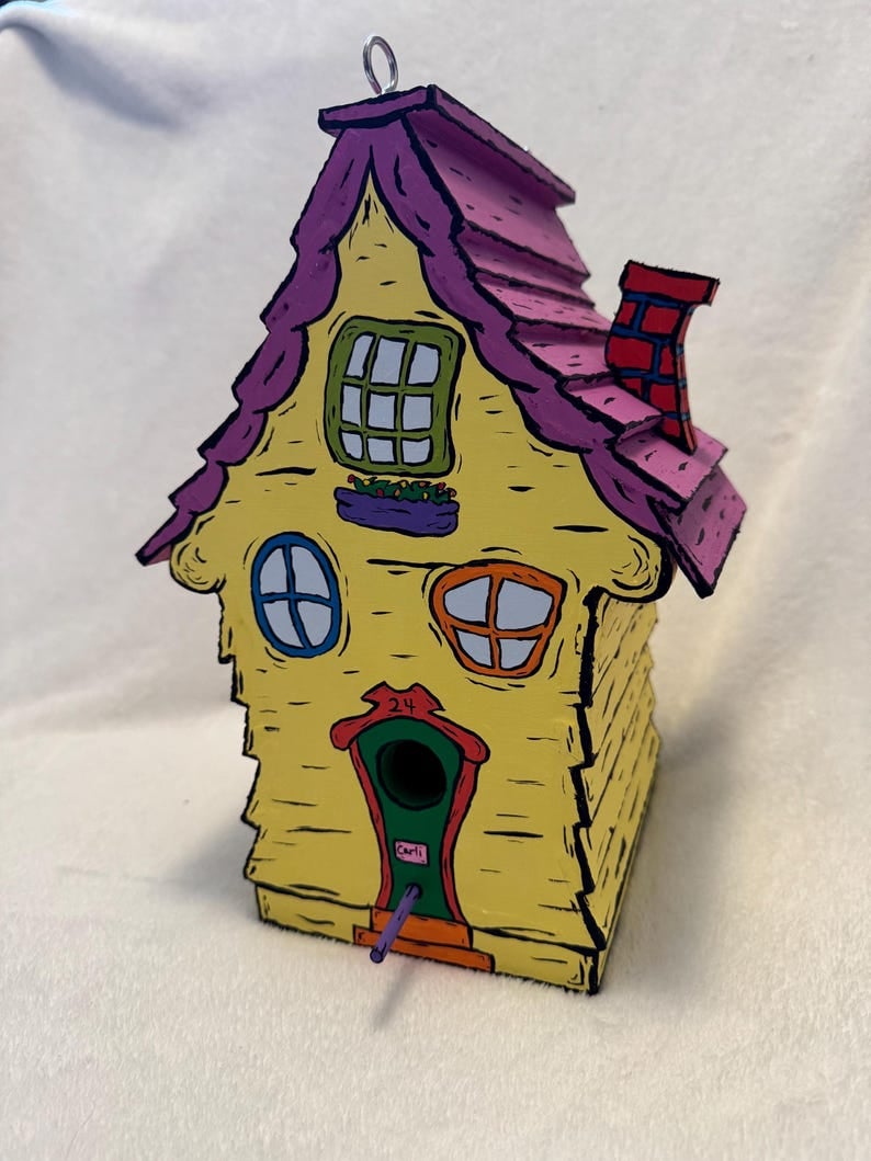 May include: A yellow birdhouse with a pink roof and a red chimney. The birdhouse has a green door with the word 'Carli' written on it. The birdhouse has a purple hanging loop and a purple dowel rod sticking out of the bottom. The birdhouse has a number '24' above the door.