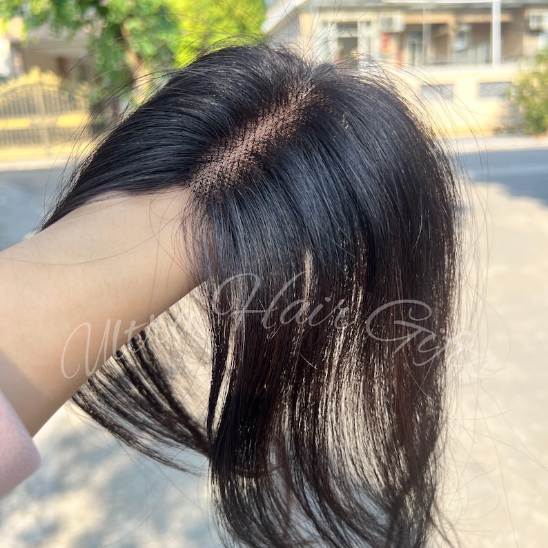 May include: A close-up of a black hairpiece with a lace front. The hairpiece has a natural-looking hairline and is held up by a hand. The text "Ultimate Hair Girl" is visible in the image.