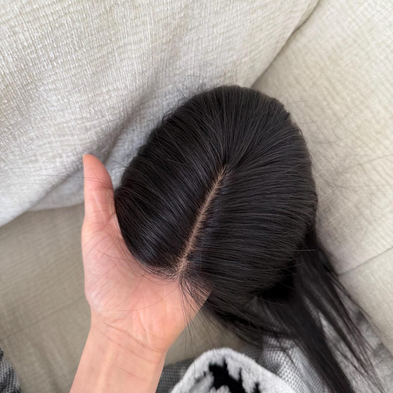 May include: A dark brown wig with a center part. The wig is held in a hand, showing the natural-looking hairline and the texture of the hair. The hair is sleek and appears to be straight.