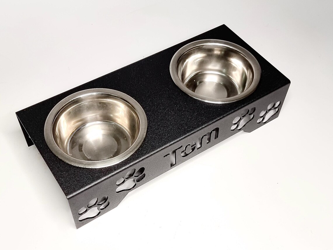 May include: Black metal double dog bowl with stainless steel bowls.  The feeder has cut-out paw prints and the name 'Tom' is engraved.  This pet feeder is ideal for small to medium-sized dogs.
