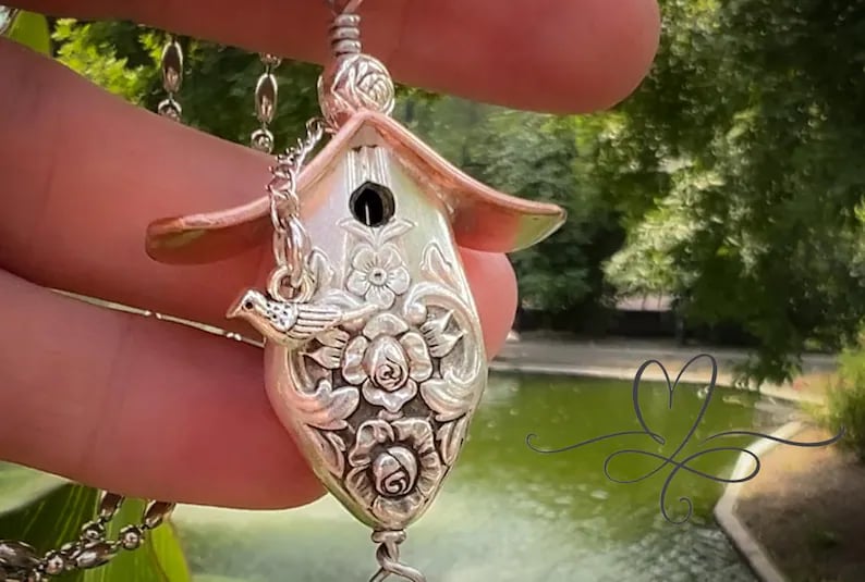May include: A silver birdhouse pendant necklace with a detailed floral design. The birdhouse has a small opening for a bird to enter and a small bird perched on the side. The necklace is on a silver chain.