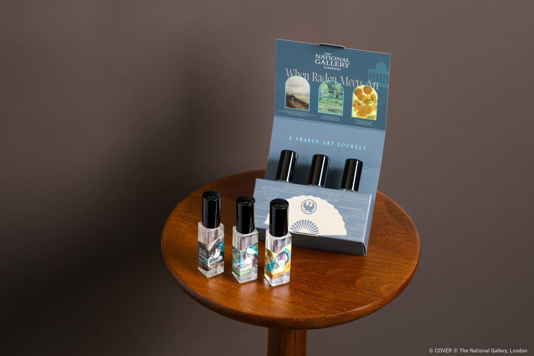 Three small fragrance mist bottles with Raden and painting-themed labels standing in front of a light blue When Raden Meets Art display box on a round wooden table