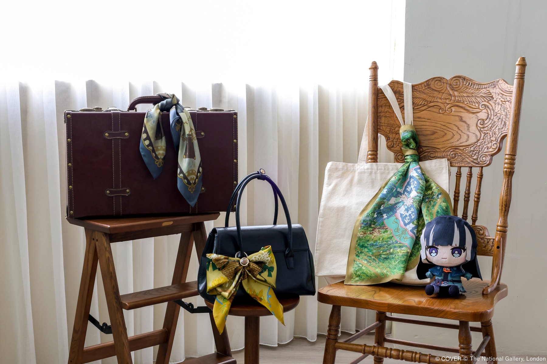 Lifestyle photo of three painting-themed silk scarves tied to a handbag and a suitcase and draped over a wooden chair beside a tote bag in soft window light