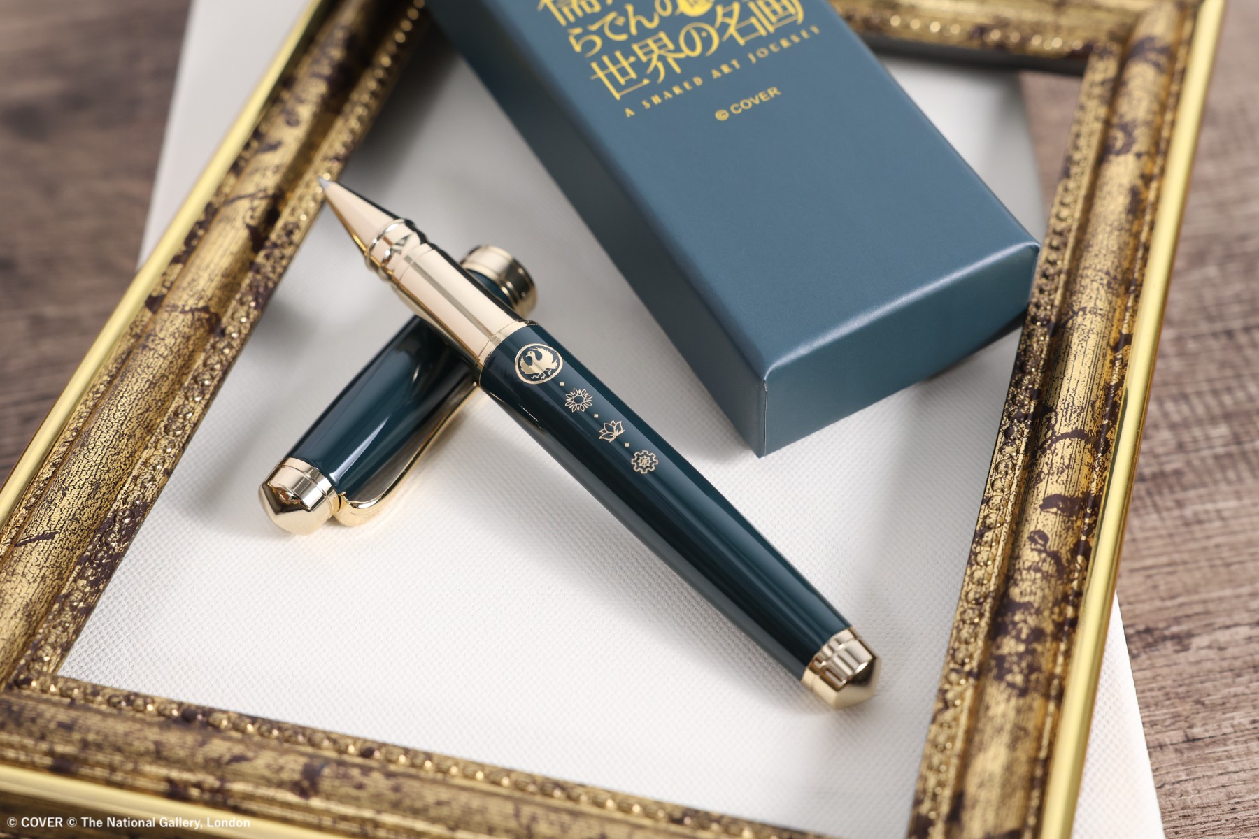Dark teal rollerball pen with gold-coloured trim and its matching presentation box resting inside an ornate gold picture frame
