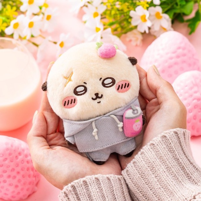 Sweatshirt-style Rakko mascot cupped in hands against a pink strawberry-milk backdrop