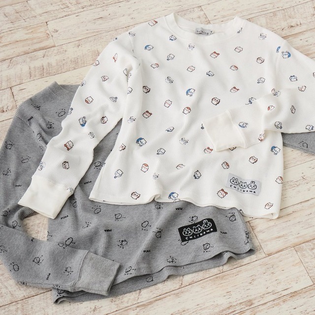 Two Chiikawa waffle long-sleeve tees, white and grey, with all-over character face prints laid on a wooden floor