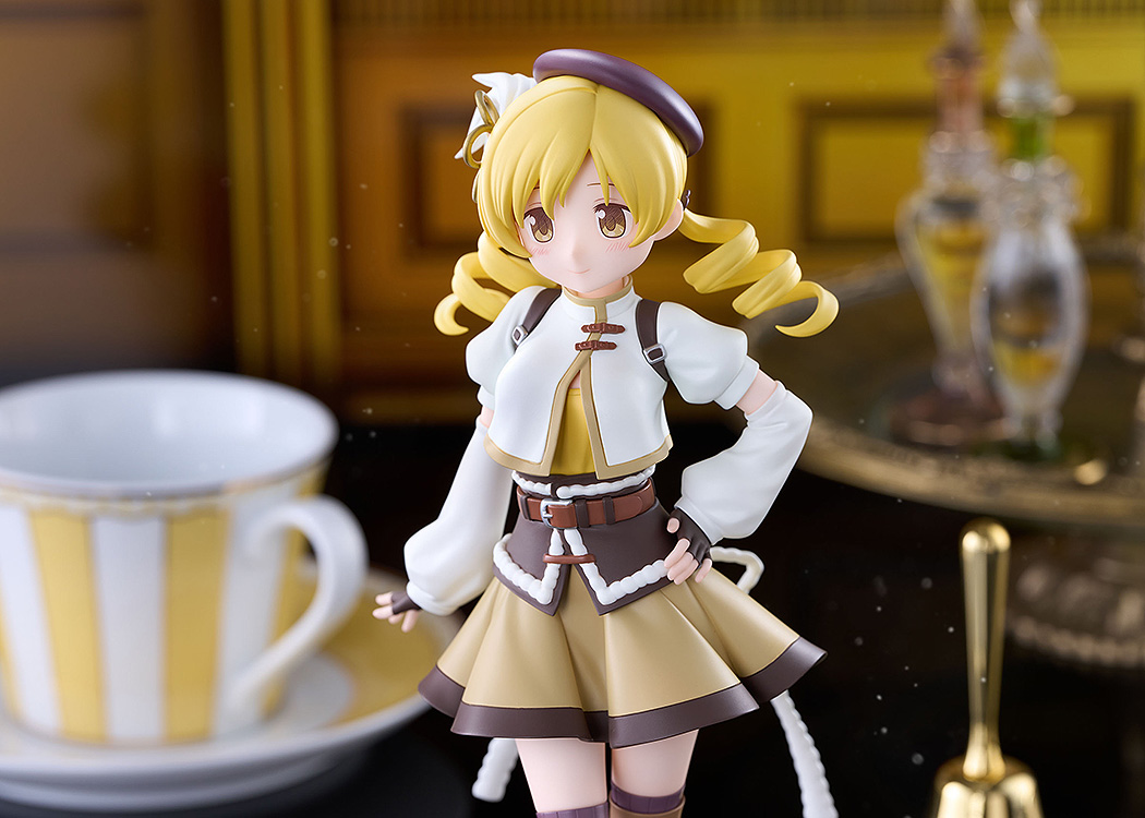 The Tomoe Mami figure posed beside a large yellow-striped teacup and a brass bell in a warm golden setting