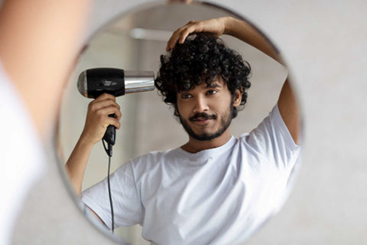 Young stylish man with black curly hair, using a blow-dryer in front of a mirror