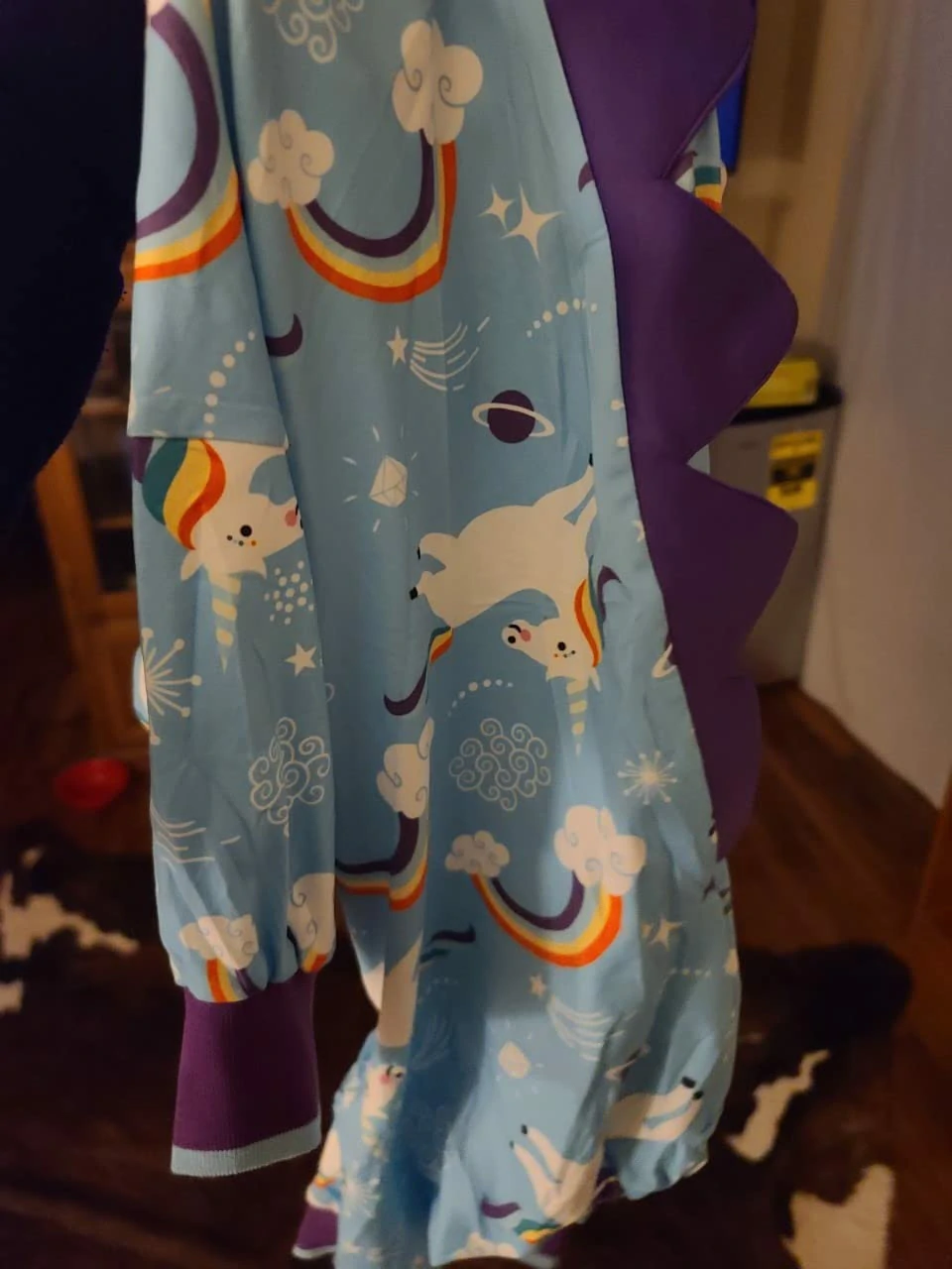 Close-up of blue dog pajamas with a unicorn and rainbow pattern, featuring purple spikes along the back, hanging indoors.