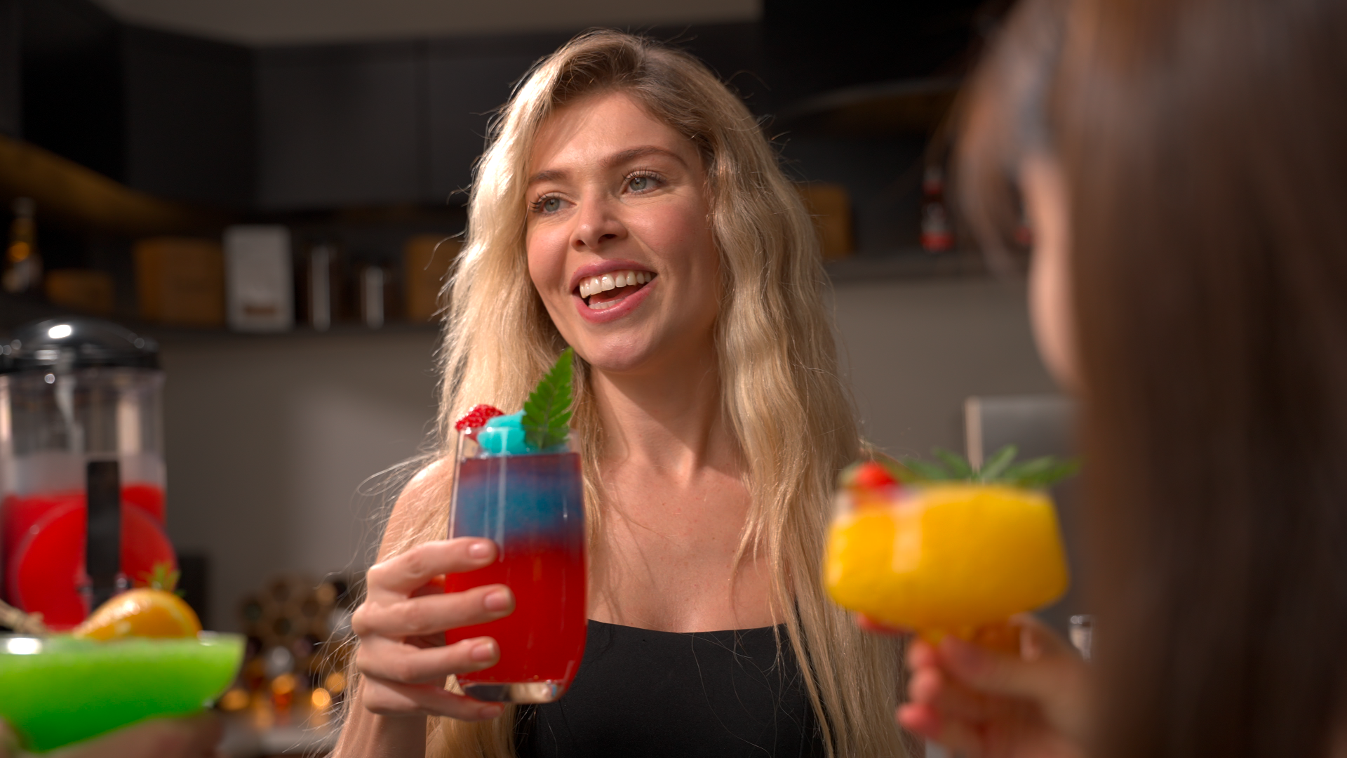 A woman smiling and holding a colorful slushy drink with blue and red layers, surrounded by friends holding other slushy drinks. In the background, a slushy machine is visible, suggesting a fun and vibrant setting.