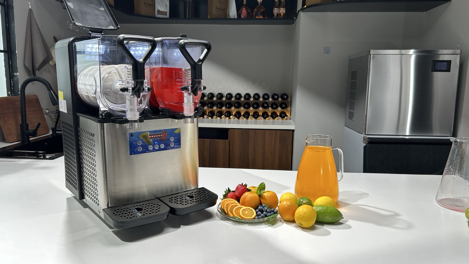 Mojgar commercial slushy machine with two flavors, surrounded by fresh fruits like oranges, lemons, and strawberries, with a pitcher of orange juice on the table in a modern kitchen setup.