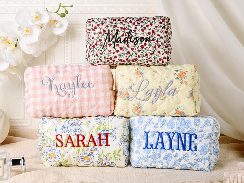 May include: Five rectangular quilted pouches with zippers, each featuring a unique fabric pattern and embroidered name. The top pouch has a red floral print with the name Madison. Below are a pink gingham pouch labeled Kaylee and a yellow floral pouch labeled Layla. The bottom row features a white pouch with blue and yellow flowers labeled SARAH and a white pouch with blue floral patterns labeled LAYNE.