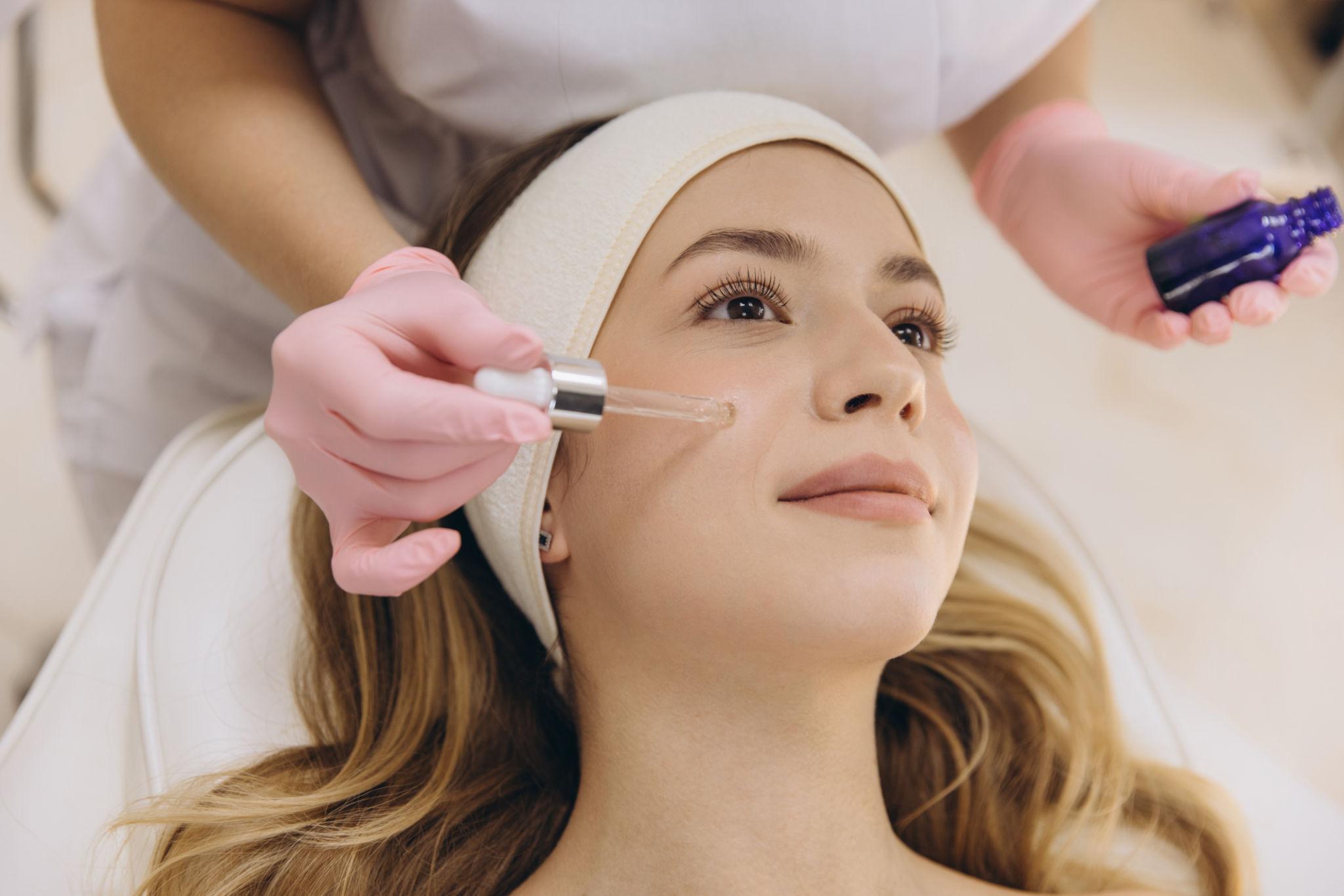 Beautician in pink gloves applying hyaluronic acid serum on blonde client receiving facial treatment at modern skincare clinic