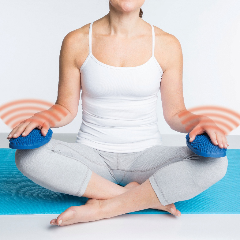 Dr Cohen’s Heatable AcuPads in use, person seated cross-legged on a yoga mat holding two blue textured heat therapy pads.