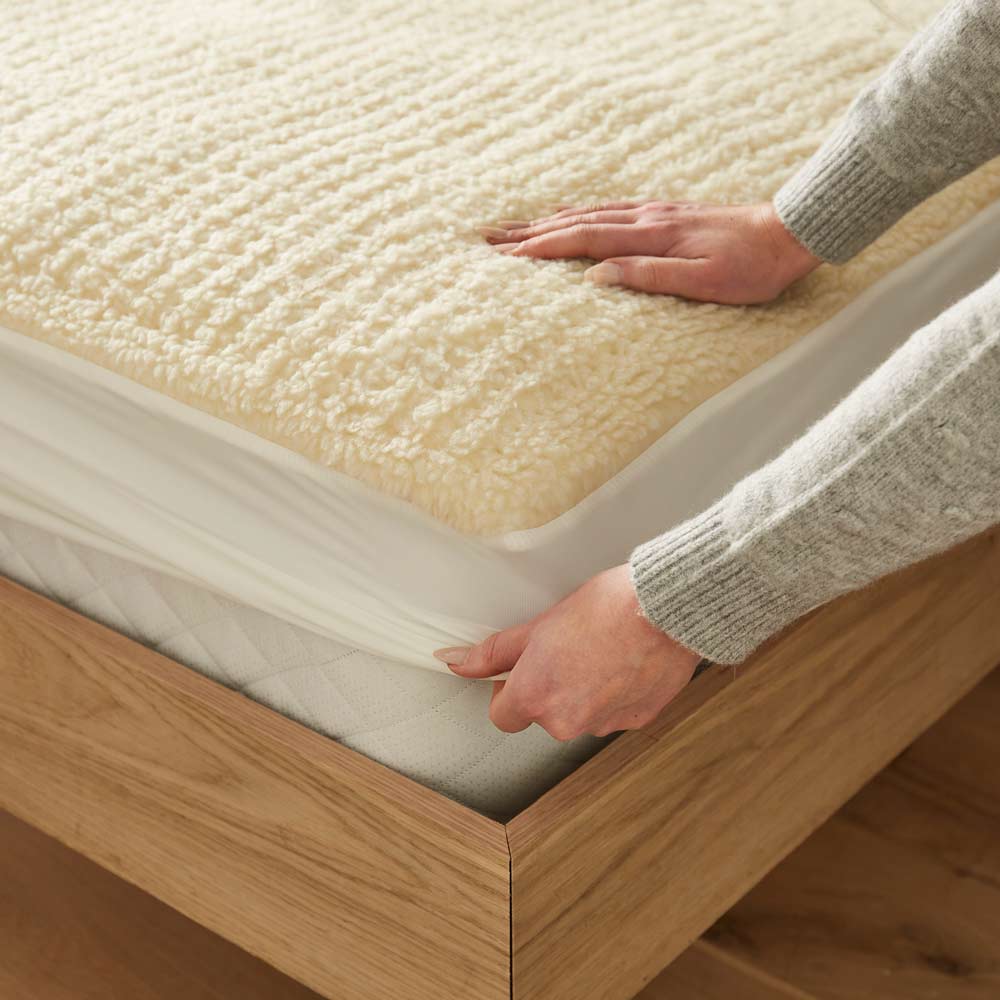 Hands fitting a Beurer heated mattress cover with deep elastic skirt onto a bed for a secure, snug fit.