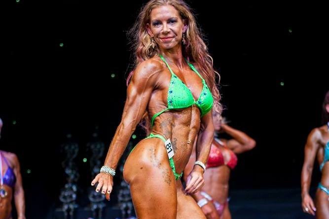 Bodybuilder, Nurse, Mum – Joanne Jagger Does It all, and We’ve Found Out How!-StressNoMore. Female bodybuilder posing on stage in a green bikini during a fitness competition, representing Joanne Jagger’s strength and athletic achievements.