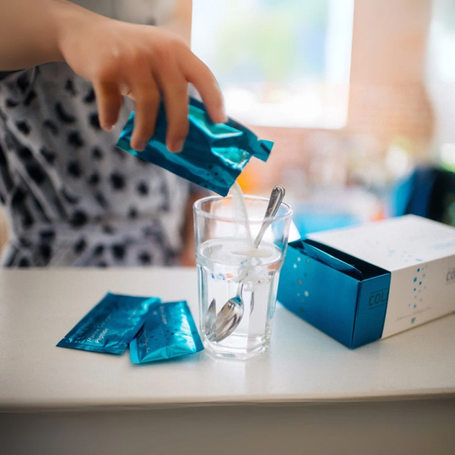 Person pouring Collamedic Bioactive Marine Collagen sachet into a glass of water, showing how to prepare the daily collagen drink for skin hydration, elasticity, and beauty-from-within support, with sachets and box visible on table.
