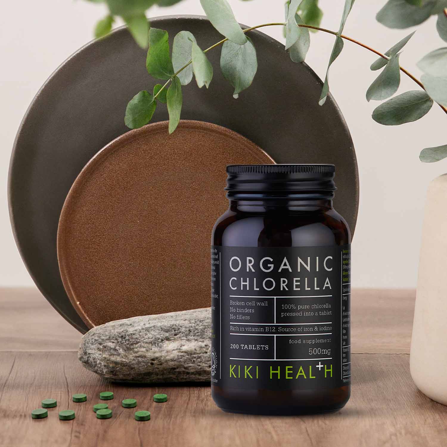 KIKI Health Organic Chlorella Tablets in a dark glass jar displayed on a wooden surface with green tablets, natural plates, stone, and foliage in the background.