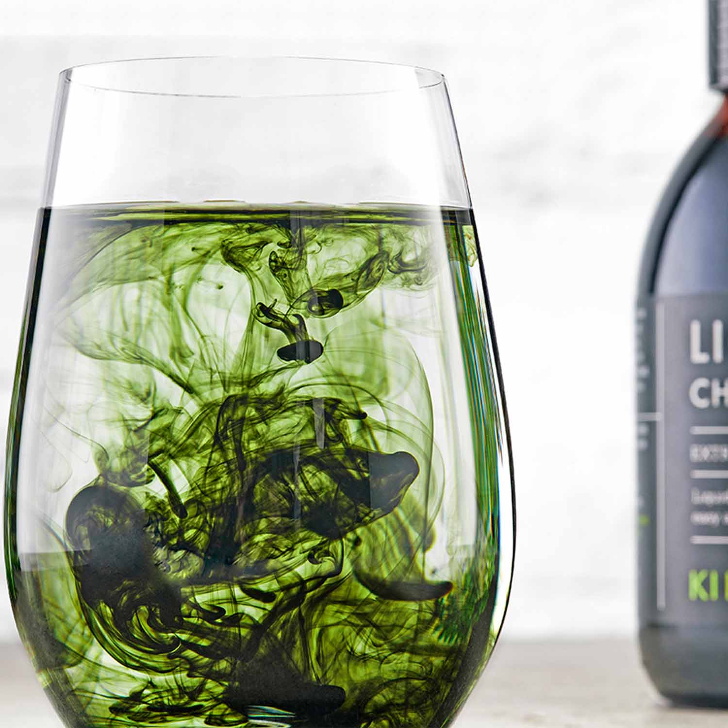 Liquid Chlorophyll 125ml - Glass of water with liquid chlorophyll dispersing into swirling green patterns, illustrating how chlorophyll mixes easily for a detox drink, with a KIKI Health Liquid Chlorophyll bottle blurred in the background.
