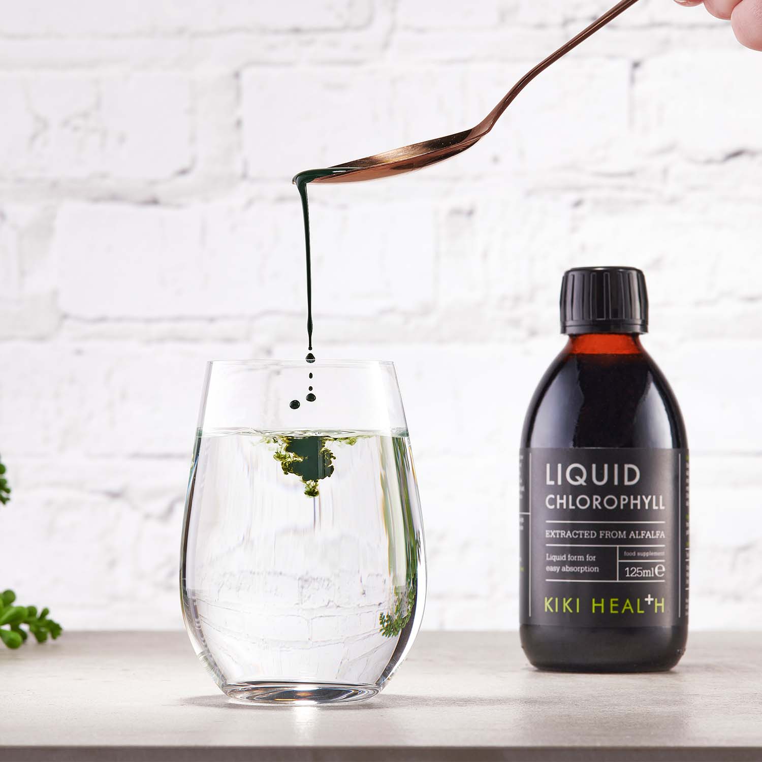 Spoon adding liquid chlorophyll drops into a glass of water, showing green chlorophyll dispersing for a detox drink, with KIKI Health Liquid Chlorophyll bottle displayed on a kitchen surface in the background.