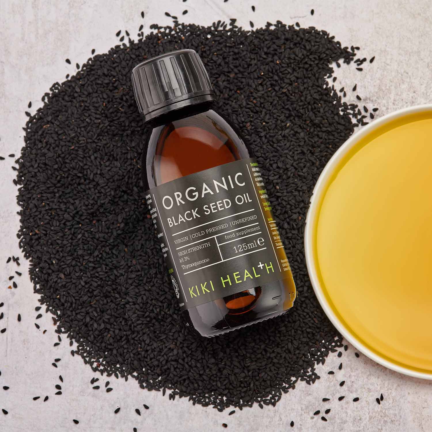 KIKI Health Organic Black Seed Oil bottle placed on a bed of black seeds beside a bowl of golden oil, photographed from above.