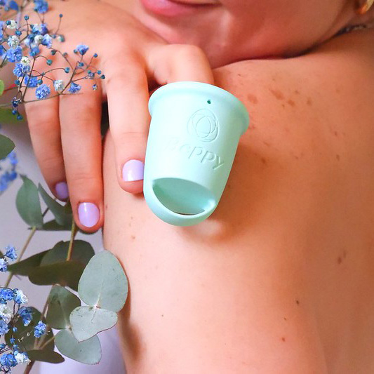 Person holding a mint green Beppy reusable menstrual cup against their shoulder, shown with natural flowers in a soft lifestyle setting.