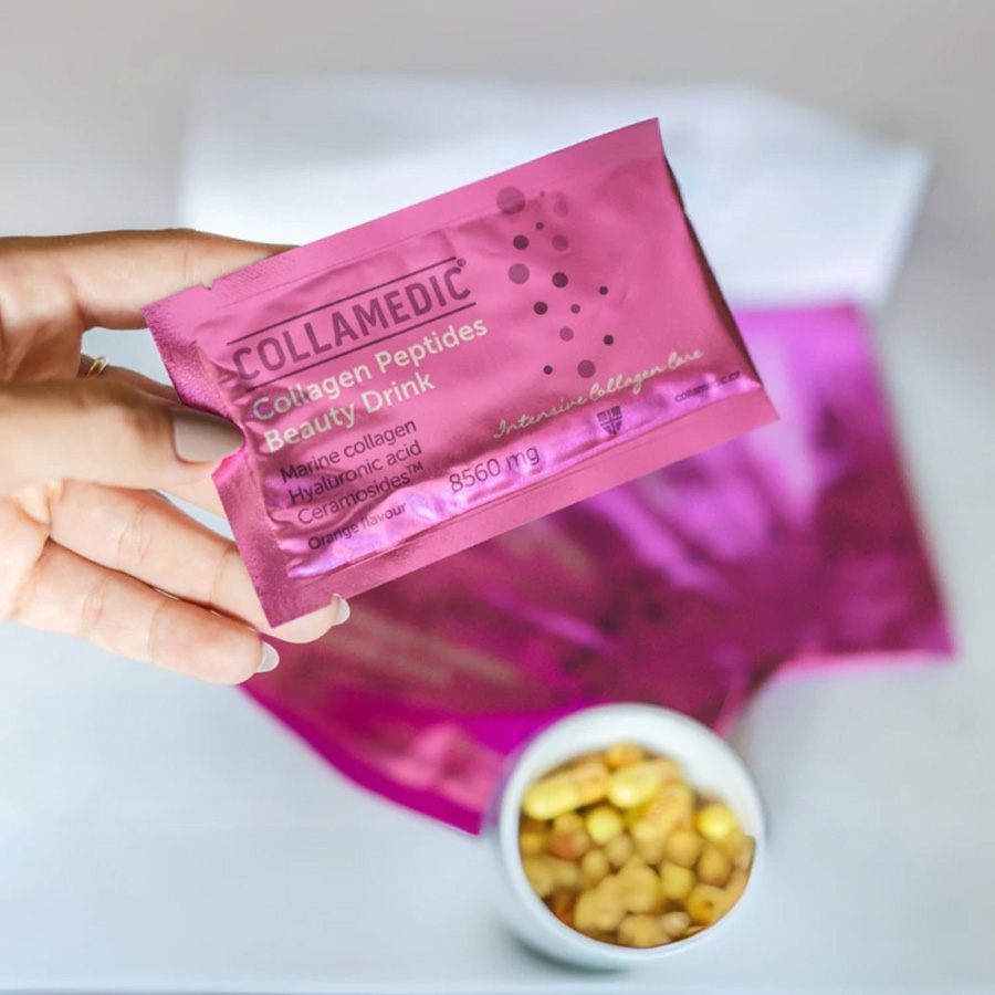 Hand holding COLLAMEDIC Collagen Peptides Beauty Drink sachet in pink packaging, showing single-serve marine collagen drink with hyaluronic acid and ceramides, with additional sachets and supplements visible in the background.