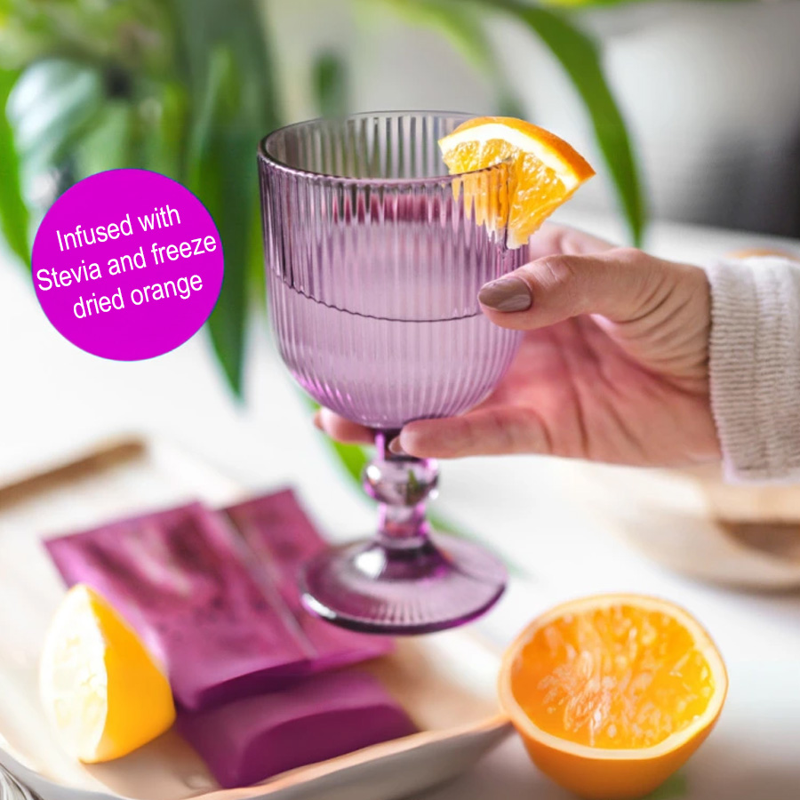 Glass of COLLAMEDIC Collagen Peptides Beauty Drink held by hand, garnished with orange slice, showing prepared marine collagen drink infused with stevia and freeze-dried orange, with sachets and fresh fruit displayed nearby.