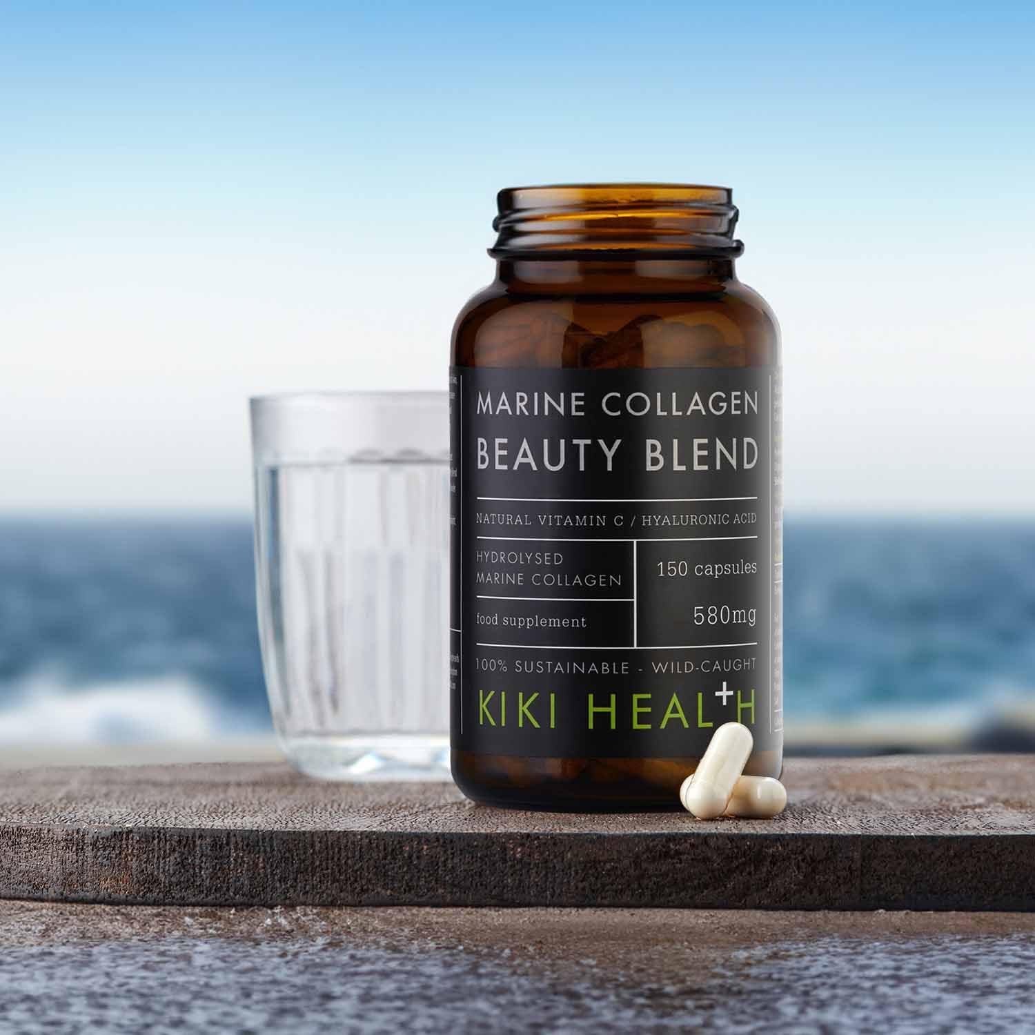 KIKI Health Marine Collagen Beauty Blend capsules in a dark glass jar displayed on a stone surface with a glass of water and capsules beside it, set against a sea background.