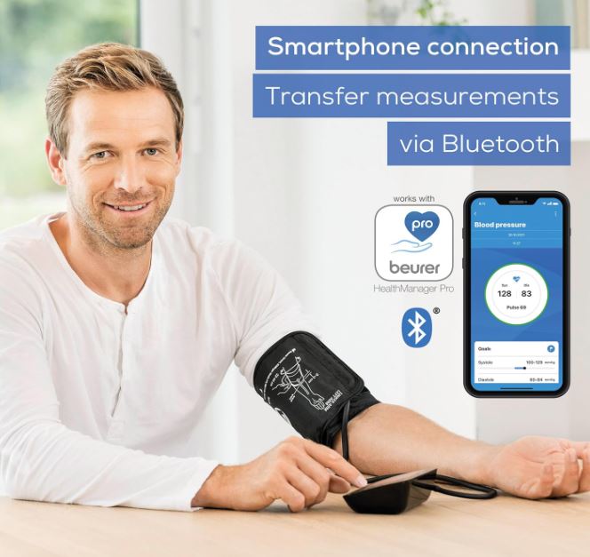 Man using Beurer BM54 upper arm blood pressure monitor with universal cuff while transferring readings via Bluetooth to a smartphone using the Beurer HealthManager Pro app for easy blood pressure tracking at home.