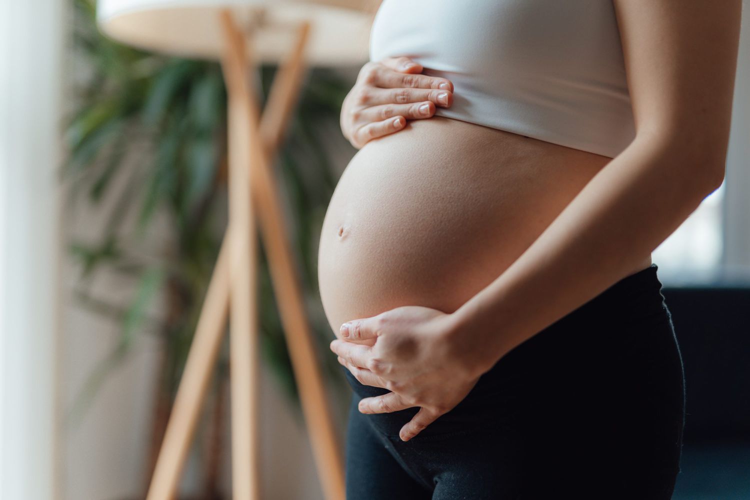 Using Vibrators During Pregnancy - The Benefits of Being Intimate-StressNoMore. Pregnant woman gently holding her bare baby bump while standing indoors, showing a close-up maternity scene in natural light.
