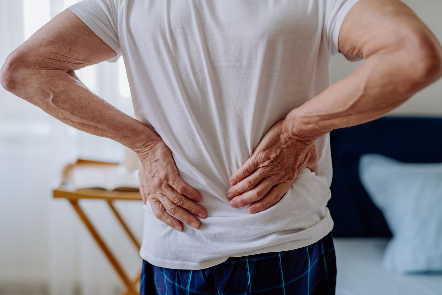 Person standing indoors with hands placed on their lower back, showing discomfort or stiffness, used to illustrate the need for support or techniques to help relieve back pain. Need Help To Relieve Back Pain?-StressNoMore