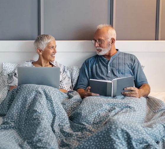 Senior couple in bed using a laptop and reading a book, showing comfort and relaxation at home. Ideal lifestyle imagery for wellness, home comfort, retirement living, or bedroom products.