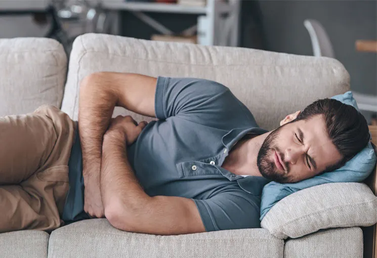 Man lying curled on a sofa holding his lower abdomen in visible discomfort, illustrating pelvic or urinary pain often associated with conditions like prostatitis. Painful Penis? Get Help for Prostatitis!-StressNoMore