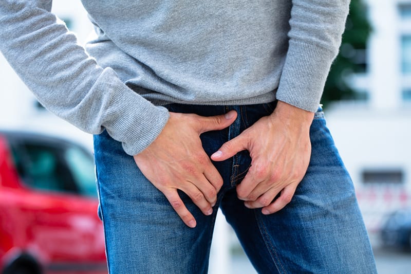 Man outdoors holding his groin area over his jeans, showing discomfort or urgency often linked to male incontinence, used to illustrate topics such as the effects of winter on bladder symptoms. The Weird Truth About Winter & Male Incontinence-StressNoMore