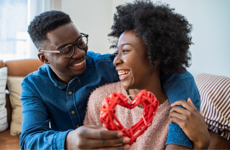How To Kick Self-doubt and Find Self-love This Valentine’s Day-StressNoMore. Smiling couple sitting together at home, holding a red heart-shaped decoration and sharing a joyful, affectionate moment