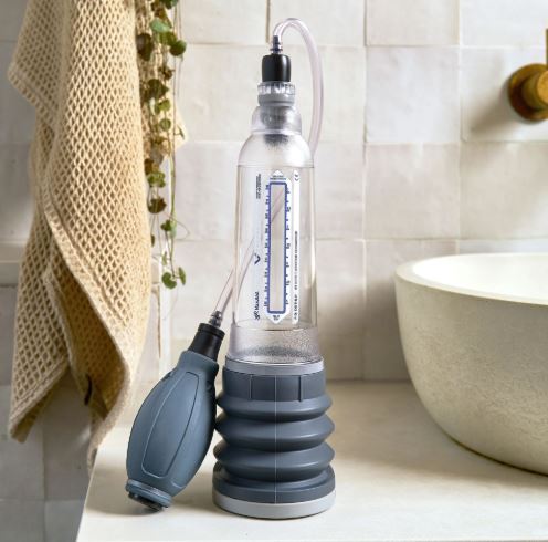 VaxAid v7 Deluxe hydro penis pump standing in a bathroom setting, showing vacuum cylinder with pressure gauge, handball pump, tubing, and soft comfort base, designed for safe erectile dysfunction treatment using water or air.