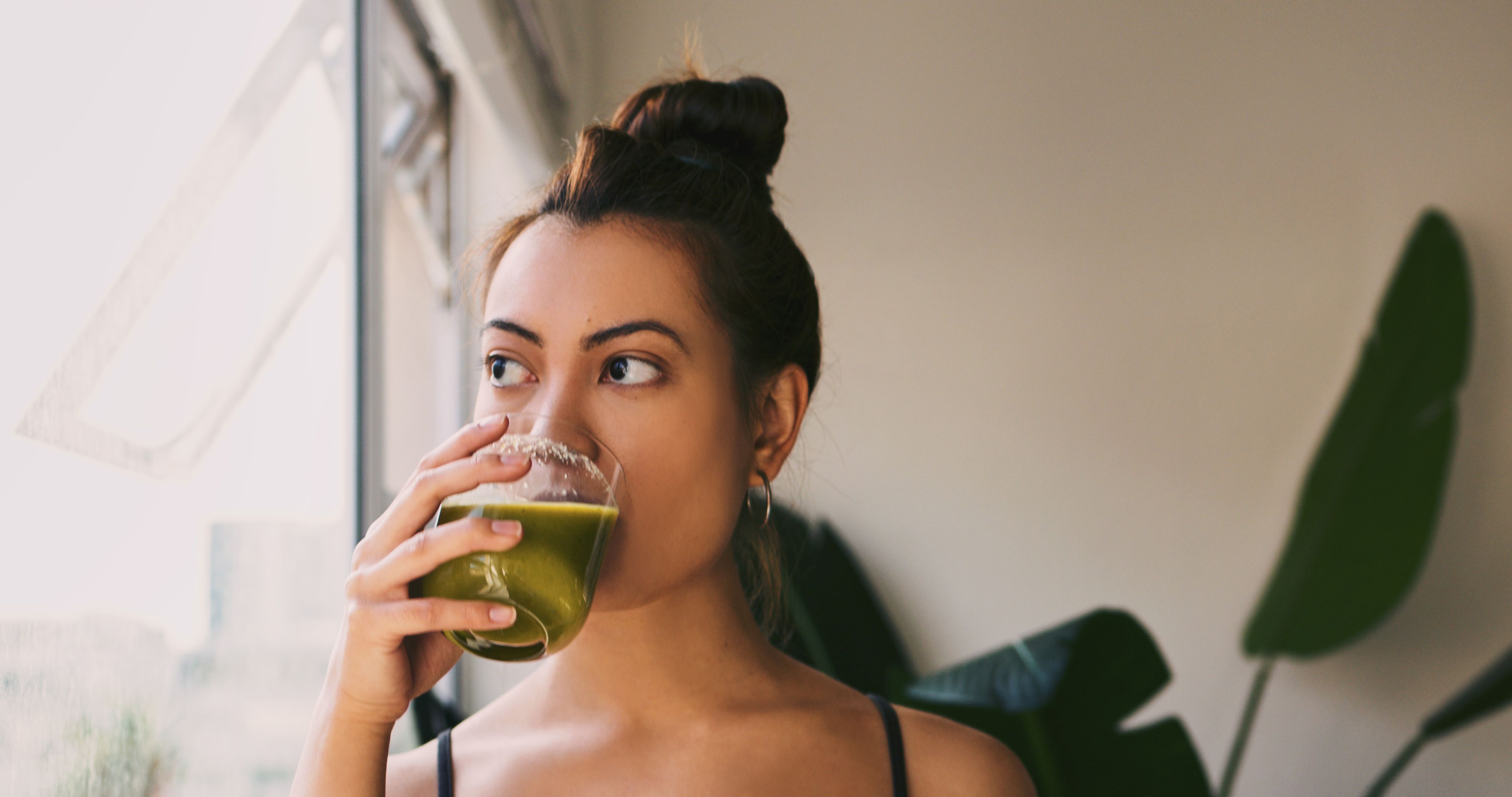 Why You Feel Rubbish During a Detox-StressNoMore. Woman drinking a green detox smoothie while looking out of a window, representing common feelings of fatigue or discomfort during a detox.