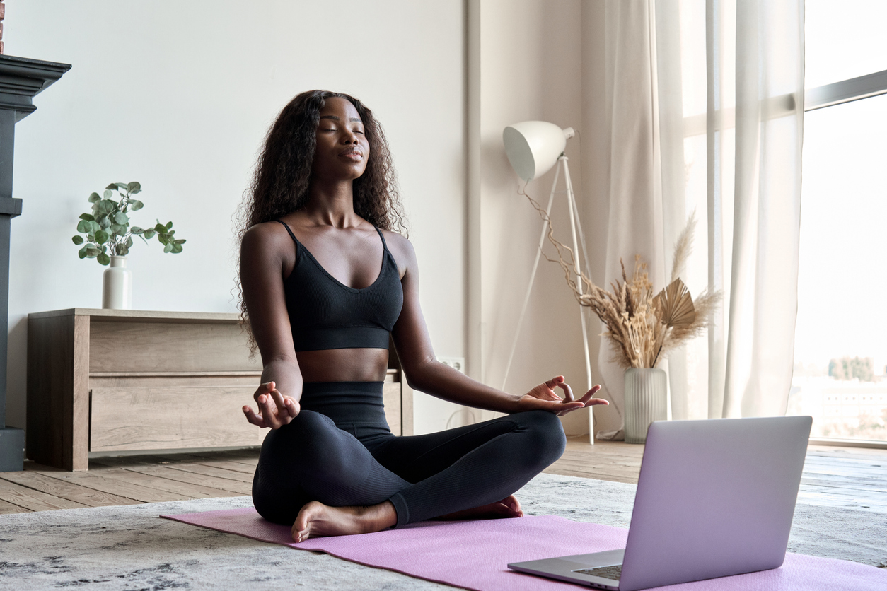 Science-Backed Benefits Of Meditation Practices For All Ages-StressNoMore. Woman practising yoga and meditation at home on a mat while following a laptop session, representing mindfulness, stress relief, home workouts and wellbeing routines.