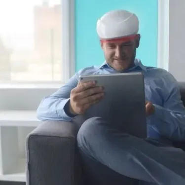 Person sitting on a sofa using a tablet while wearing a Collagenius Red light therapy helmet designed for hair regrowth. Low-Level Laser Therapy (LLLT) for Hair Loss. Stressnomore.
